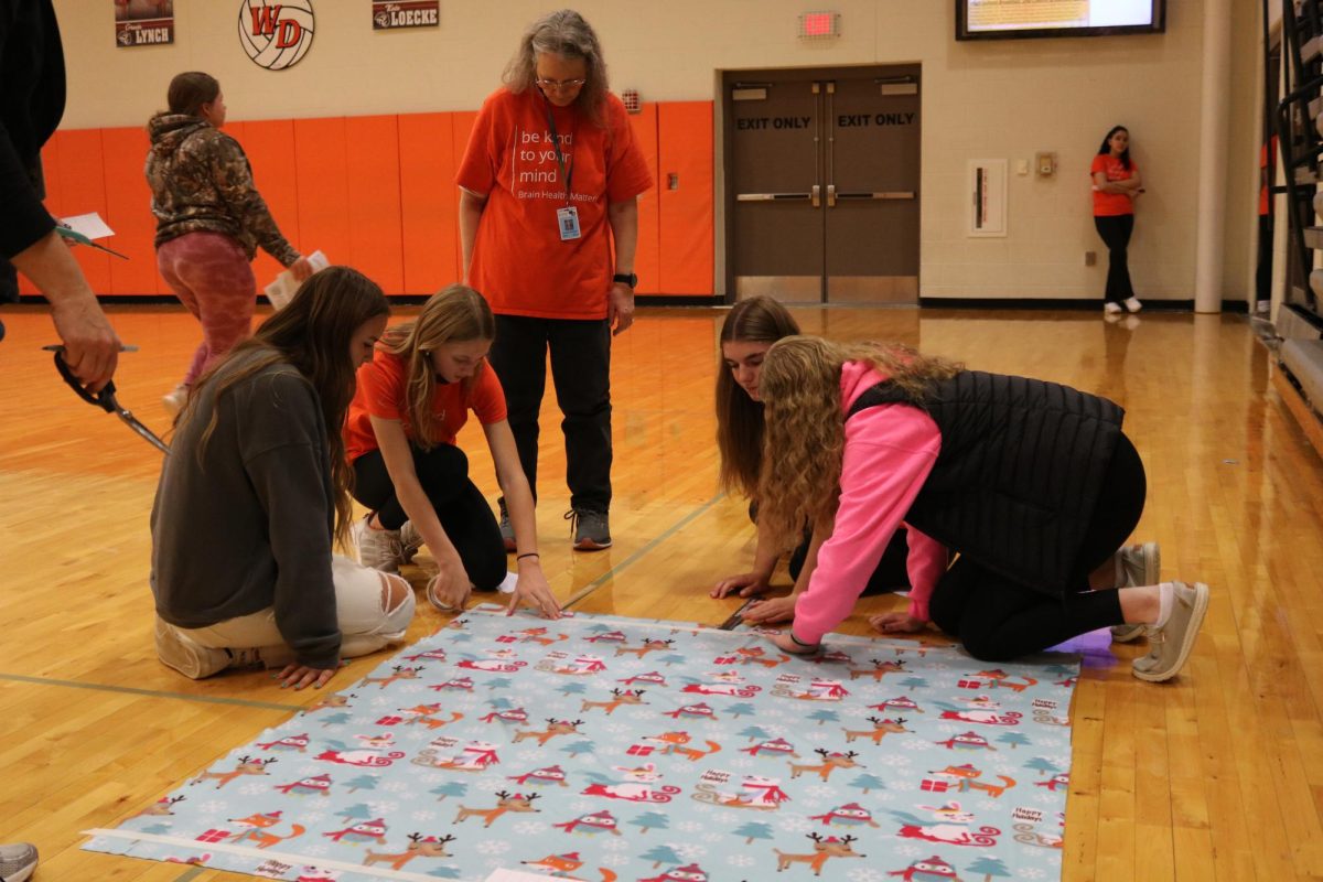 Adler Mormann (9), Avery Schmitz (9), Parker Robinson (9), and Kate Ries (9) participate in cutting tie blankets for project Linus as teacher Marianne Sandberg looks on. 