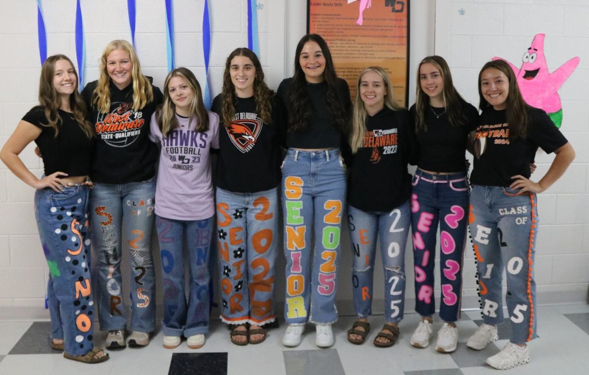 Seniors pose wearing their senior jeans: Sydney Shover, Paige Woellert, Lili Shappell, Amelia Schnieders, Andrea Wubbena, Kenzi Hempstead, Andrea Kann, and Addison Burger. Seniors used paint along with glitter, ribbons, and other materials to decorate their jeans. 